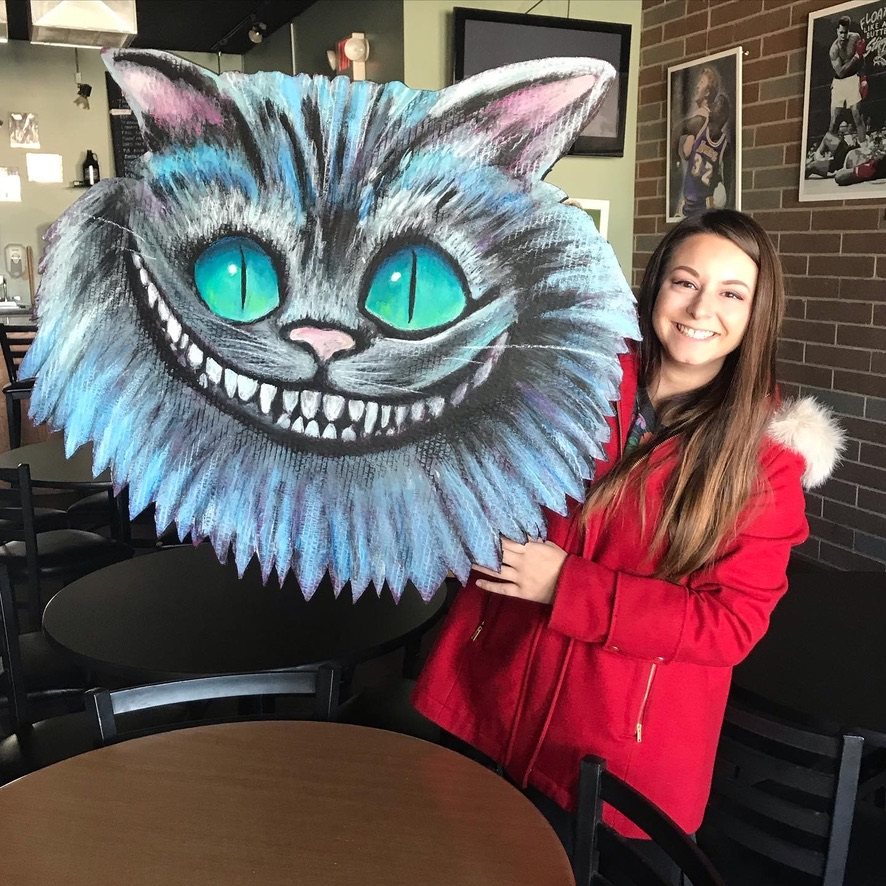 Hand-cut and hand-painted Cheshire Cat face in blue and gray with teal eyes and a wide grin, held by the customer in a restaurant