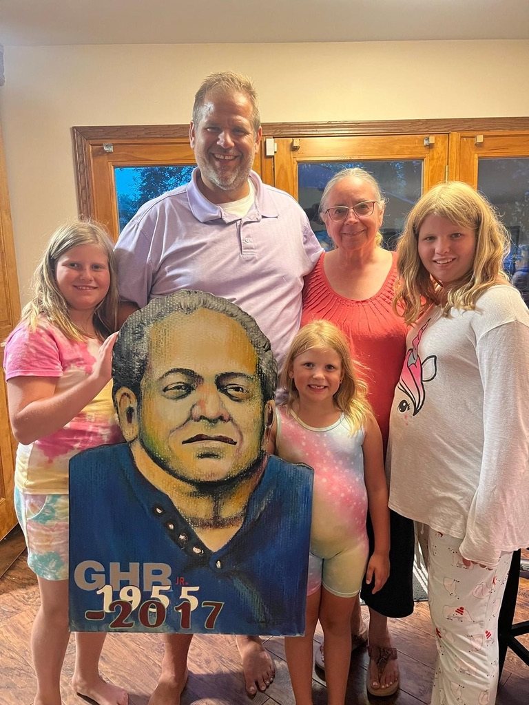 Hand-cut and hand-painted memorial portrait of a man, lettered GHR Jr. 1955 to 2017, held by his family in their home