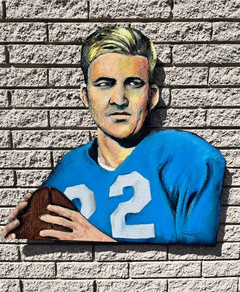 Hand-cut and hand-painted portrait of a blond Detroit Lions quarterback, number 22, in a blue jersey holding a football, on a brick wall