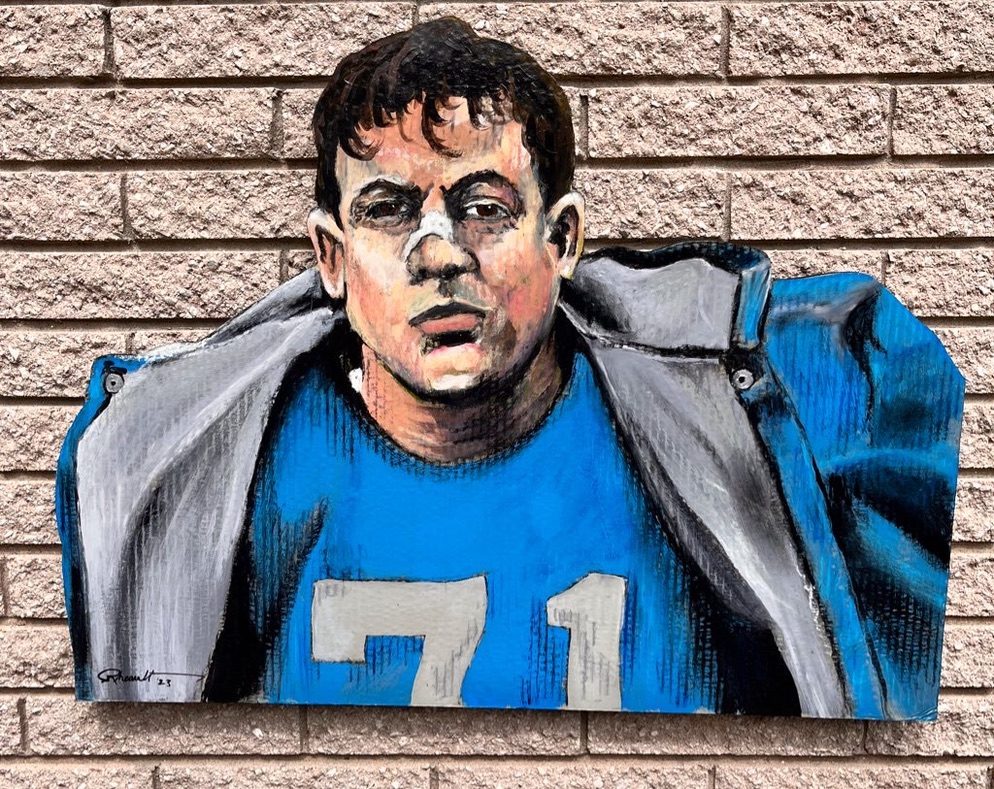 Hand-cut and hand-painted portrait of a Detroit Lions player, number 71, in a blue jersey under a gray jacket, on a brick wall