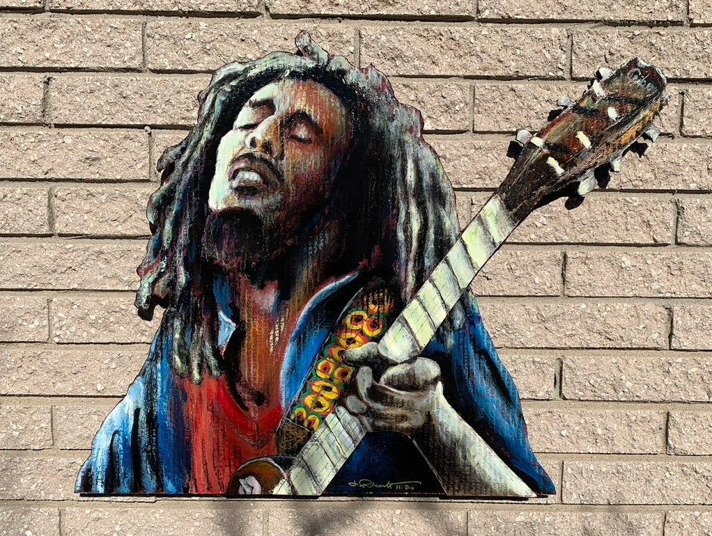 Hand-cut and hand-painted portrait of Bob Marley playing an acoustic guitar, on a brick wall